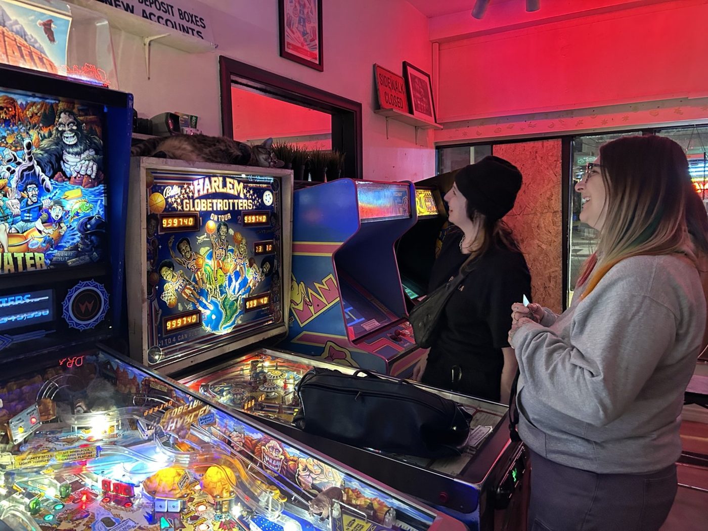 Two women laughing beside vintage pinball machines under red light